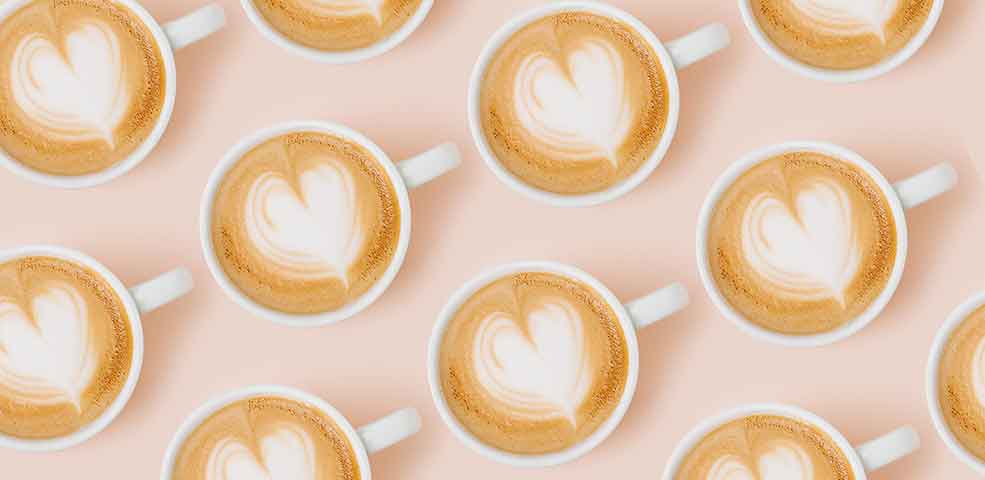 Multiple coffee mugs with latte art hearts in each and made with the best organic espresso. Shown on a peach background.