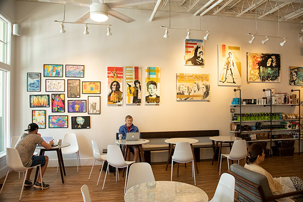 Coffee Shop with people sitting at tables. The image is focused on a wall with paintings all down it.