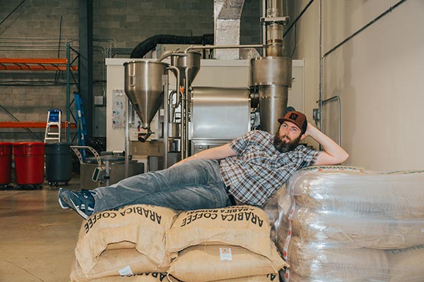Colt at lounging with stacked burlap bags of green coffee beans at Amavida Coffee Roasters. Colt is the Operations Manager at Amavida, leading all the wholesale production within our roaster and distribution center, while also being a part of serving on committees with Cooperative Coffees.